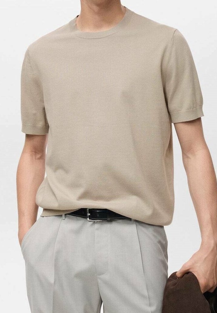 Homme portant un haut en maille beige à manches courtes rentré dans un pantalon plissé gris clair avec une ceinture noire, tenant une veste marron foncé.