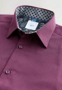 Burgunderfarbene Button-up-Bluse aus strukturiertem Stoff, mit einem gemusterten Innenkragen, der florale und geometrische Designs aufweist. Markenlabel sichtbar.
