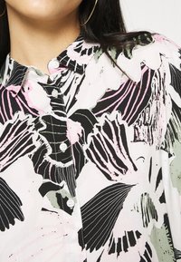 Blouse fleurie présentant un fond blanc avec des motifs botaniques en noir et rose audacieux, un col classique et une fermeture à boutons sur le devant.