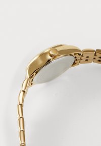 Montre-bracelet dorée avec un boîtier poli, un cadran rond, une couronne proéminente et un bracelet à maillons. Texture lisse avec une finition réfléchissante.