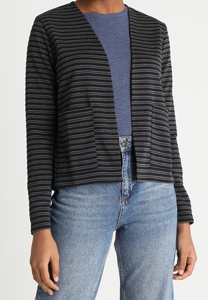 Vrouw draagt een zwart-grijs gestreepte vest met open voorkant over een blauw geribd topje en lichtblauwe high-waisted jeans.