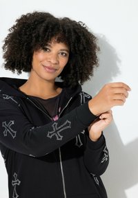 Felpa nera con zip e design di croci metalliche, polsini in costina e una texture morbida. Il modello mostra il dettaglio delle maniche su uno sfondo bianco.