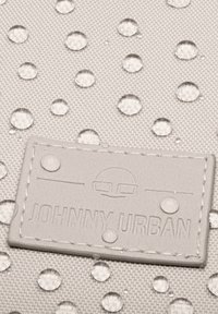 Szürke szövet vízcseppekkel, amelyen egy bőrcímke látható, rajta a "JOHNNY URBAN" felirattal és egy logóval felette.