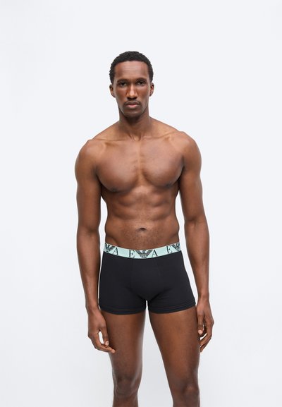 Μυώδης άνδρας στέκεται, φορώντας μαύρα εσώρουχα μποξέρ με ανοιχτό μπλε ζώνη στη μέση που φέρει λογότυπο, σε λευκό απλό φόντο.