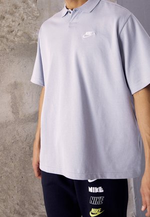 Polo gris clair avec un col classique et des manches courtes. Présente un logo Nike blanc sur la poitrine gauche, fabriqué en tissu en coton doux.