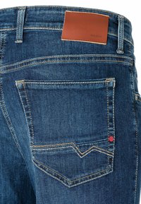 Blaue Denim-Jeans mit einem Lederpatch, der "MAC JEANS" beschriftet ist, mit kontrastierendem orangem Nähen und einem roten Logodetail auf der Tasche.