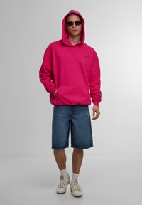 Felpa con cappuccio fucsia, dotata di tasca frontale, abbinata a pantaloni shorts di denim. Gli accessori includono occhiali da sole scuri, sneakers bianche e calze corte.