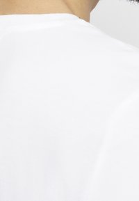 Detailní záběr na krk a rameno osoby, která má na sobě jednoduché bílé tričko s kulatým výstřihem na světlém pozadí.