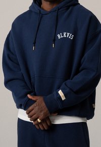 Man in een marineblauwe hoodie met de tekst "BLKVIS" en bijpassende broek, met gekruiste handen vooraan, gouden ring om de linkerste hand.