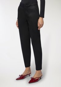 Pantaloni neri su misura con una texture liscia, dotati di una cintura e tasche laterali. Abbinati a mule rosse con punta appuntita e dettagli con fiocco.