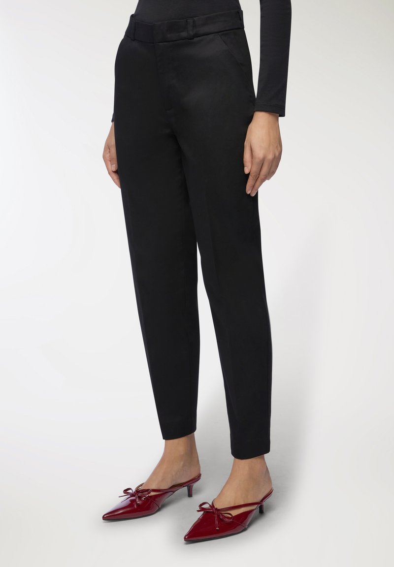 Pantaloni neri su misura con una texture liscia, dotati di una cintura e tasche laterali. Abbinati a mule rosse con punta appuntita e dettagli con fiocco.