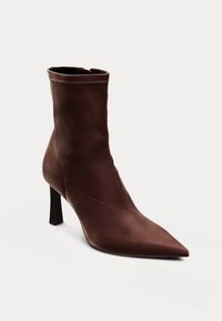 Bottine marron à bout pointu, texture lisse, talon haut fin et design minimaliste. Présente des coutures nettes et une fermeture zippée.