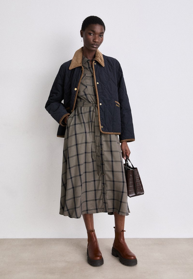 Frau in einem karierten Midikleid, mit einer marineblauen gesteppte Jacke mit hellbraunem Kragen, braunen Ankle Boots, die eine dunkelbraune Handtasche hält.
