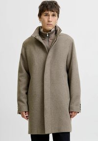 Üks suur brown villane mantel kõrge kraega, millel on peidetud kinnitused ja nööbiga mansetid. Allpool on nähtav kerge, lukuga jakk.