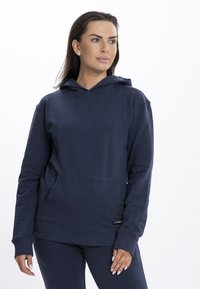 Hanorac bleumarin dintr-un material moale, având buzunar tip kangur, manșete și tiv cu pliuri, cu un etichetă logo în partea stângă jos.