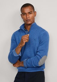 Suéter azul texturizado con un cuello alto, que presenta parches grises en los codos y un logo en el pecho. Mangas largas, superpuesto a una camisa de color azul claro.