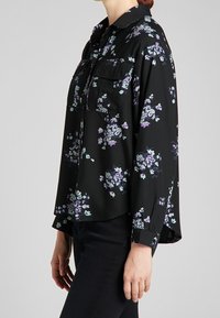 Svart blus med blommönster i lila och turkos, långärmad, knäppning fram och två bröstfickor. Slät, lätt tyg.