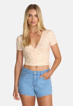 Femme blonde portant un crop top beige fleuri à nouer et un short en jean bleu clair taille haute, une main dans la poche.
