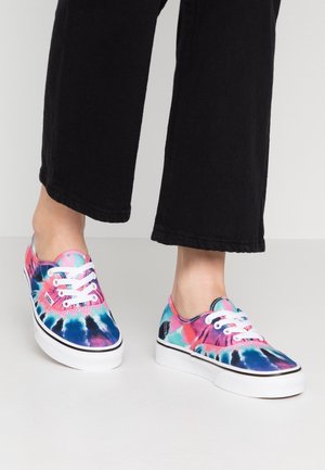 Stopy w niskich sneakerach na sznurowadła z kolorowym wzorem tie-dye w różowym, niebieskim i morskim kolorze, zestawione z czarnymi spodniami o szerokich nogawkach.