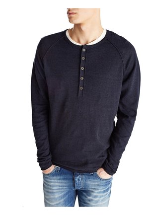 Homme portant un henley à manches longues bleu marine foncé avec cinq boutons, un sous-t-shirt blanc, et un jean bleu, les mains dans les poches.
