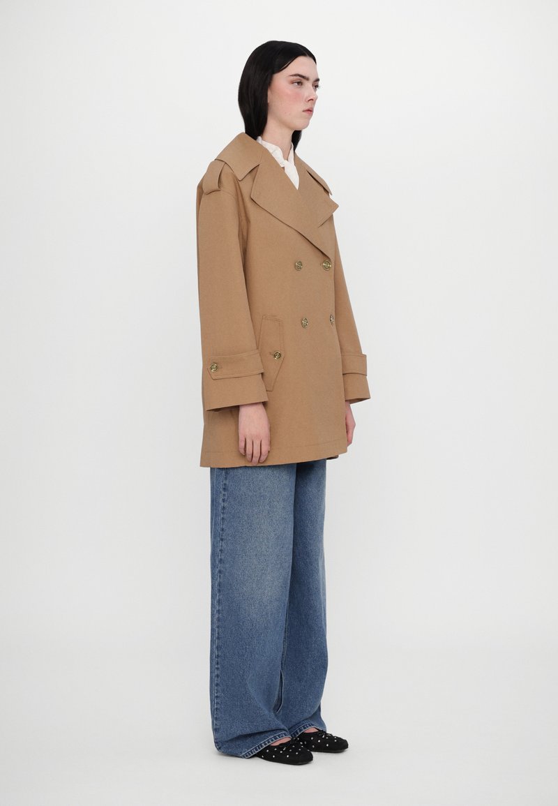 Kamelfarbiony płaszcz dwurzędowy z szerokimi klapami, złotymi guzikami i dużymi kieszeniami, w połączeniu z luźnymi niebieskimi dżinsami i czarnymi płaskimi butami.