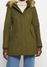 Kvinde iført en olivengrøn vinterjakke med en pelskantet hætte, beige turtleneck og blå jeans foran en ensfarvet baggrund.