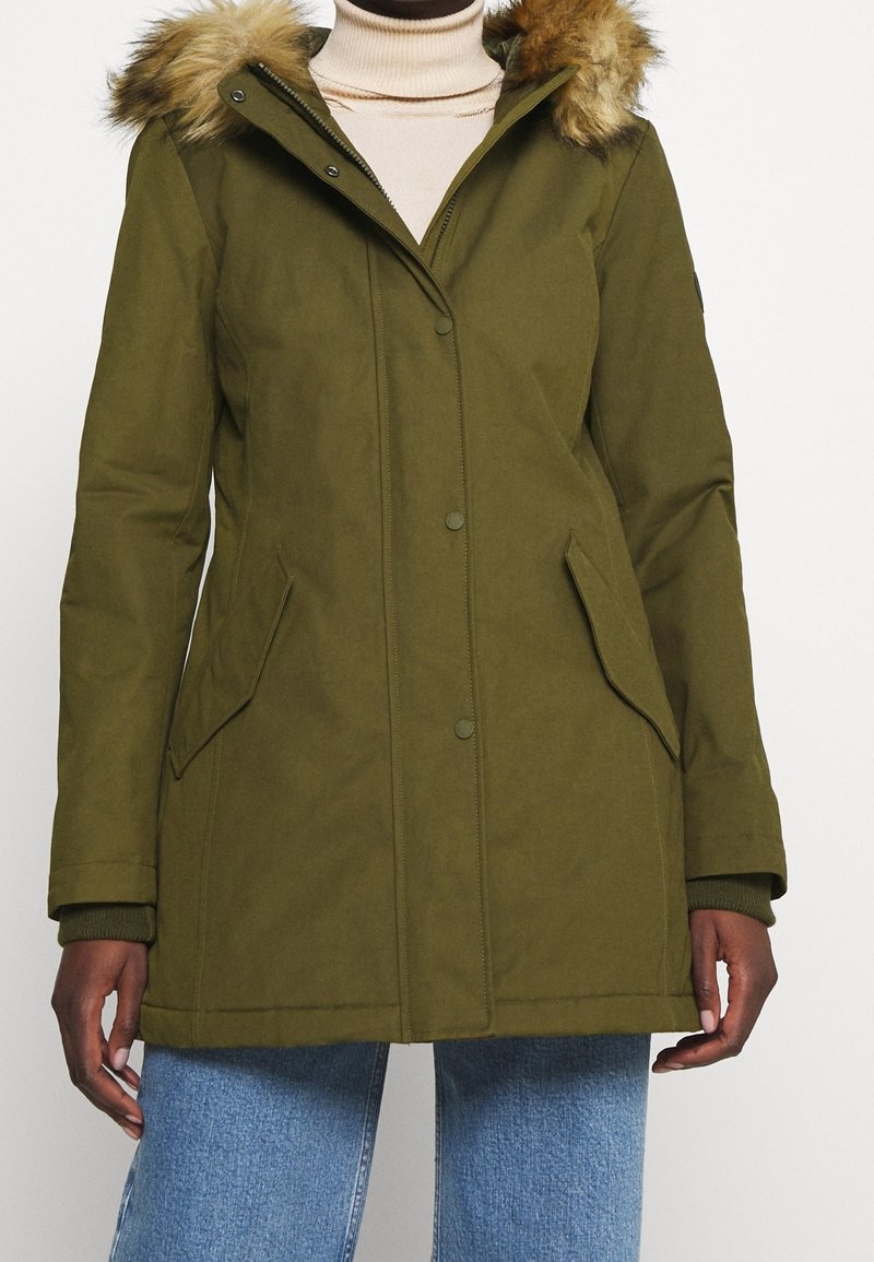 Kvinde iført en olivengrøn vinterjakke med en pelskantet hætte, beige turtleneck og blå jeans foran en ensfarvet baggrund.