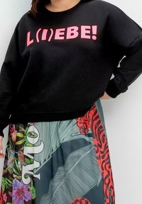 Czarna bluza z różowym napisem "L(I)EBE!" w zestawieniu z kolorową, wzorzystą plisowaną spódnicą z tropikalnymi kwiatami i liśćmi.
