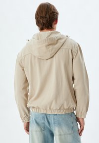 Beige huppari, jossa on elastiset hihansuut ja helma, erityisesti hieno pinta ja rento istuvuus. Selkäpuolella on ompeluyksityiskohtia ja yksinkertainen muotoilu.
