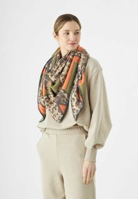 Femme vêtue d'un ensemble de détente beige avec une grande écharpe à motifs en orange, vert et marron drapée autour du cou, les mains dans les poches, regardant à droite.