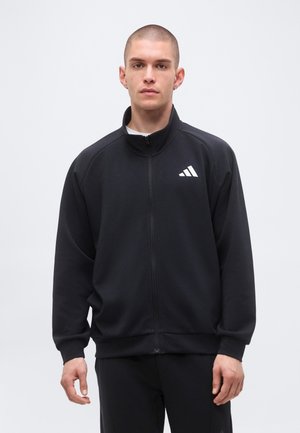 Ung mand med kort hår iført en sort Adidas jakke med hvidt logo, stående mod en almindelig lys baggrund.