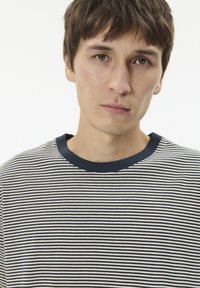 Trumpa dryžuota marškinėliai su siaurais tamsiai mėlynais ir kreminiais dryžiais. Turi vientisą tamsiai mėlyną, apvalų kaklo iškirpimą. Minkšta audinys su lygiu paviršiumi.