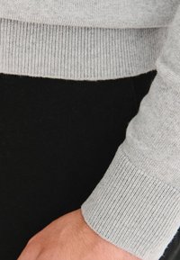 Jasnoszary sweter z dzianiny z mankietami o prążkowanej fakturze, prezentujący miękkie wykończenie i dopasowany krój. Czarna tkanina widoczna pod swetrem.