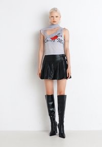 Haut sans manches gris avec un imprimé graphique, associé à une jupe plissée en simili cuir noir et des bottes à talons noires montantes.