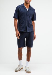 Marineblauwe short-sleeve shirt en bijpassende korte broek van textuurstof, met een knoopsluiting, platte kraag en een getailleerde pasvorm. Witte sneakers.