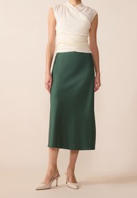 Un haut blanc sans manches froncé associé à une jupe midi satin vert foncé et des escarpins à talons hauts nude sur un fond pâle.