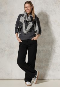Grau gestrickter Pullover, schwarze weite Hose und ein schwarz/graues gemustertes Tuch. Weiße Schuhe mit grauen Akzenten runden das Outfit ab. Neutrale Hintergrund.