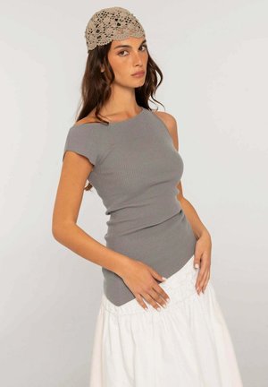 Mujer con un sombrero beige de ganchillo, top gris de un solo hombro con canalé, y una falda larga blanca de pie con las manos apoyadas en las caderas.