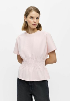 Vrouw met kort bruin haar die een lichtroze top met korte mouwen en geplooide taille en een donkere broek draagt, tegen een effen witte achtergrond.