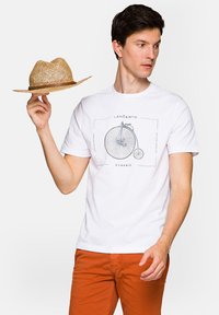 Biała bawełniana koszulka z grafiką roweru w czerni, w połączeniu z pomarańczowymi spodniami. W ręce model trzyma słomkowy kapelusz.