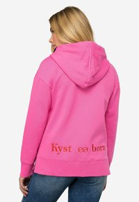 Felpa con cappuccio rosa realizzata in morbido tessuto. Presenta un ricamo arancione con la scritta "Kyst born" sul retro, con vestibilità rilassata e polsini a costine.