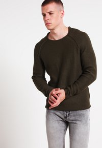 Olíva zöld texturált pulóver kerek nyakkivágással és hosszú raglán ujjal, világosszürke, megkoptatott farmernadrággal párosítva.