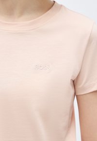 Κοντινό πλάνο ατόμου που φοράει ανοιχτό ροζ κοντομάνικη μπλούζα με την λέξη «BOSS» κεντημένη σε λευκό στο στήθος.