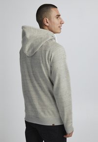Haut à capuche gris marbré avec cordon de serrage, tissu texturé, ourlet côtelé et une petite étiquette logo dans le bas du dos, associé à un pantalon foncé.