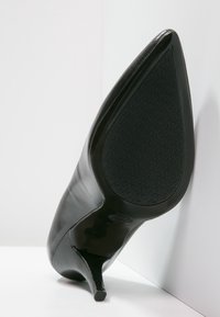 Chaussure à talon haut en cuir noir avec un bout pointu et un talon aiguille fin. La semelle présente un motif de grip texturé.