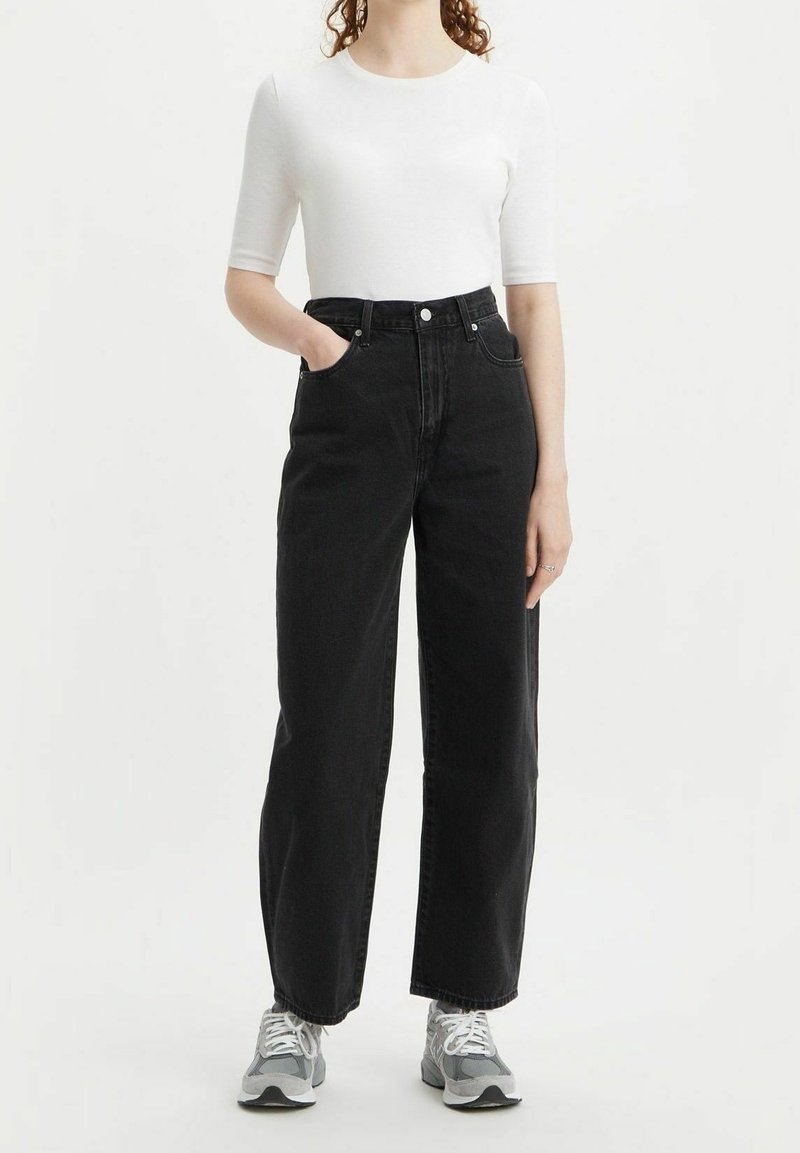 Femme portant un top blanc à manches courtes, un jean noir taille haute à jambes larges et des baskets grises, debout, une main dans la poche.