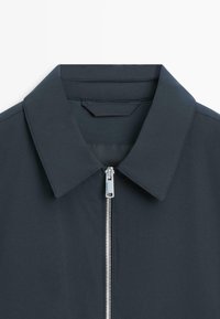 Mørkegrå jakke med krave og synlig sølvlynlås, der er helt lynet op mod en hvid baggrund.