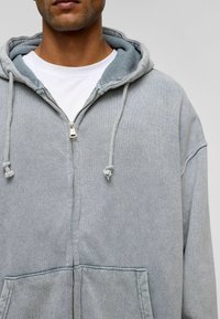 Lichtgrijze ritshoodie, gemaakt van zachte stof, met een grote capuchon, een trekkoord en voorzakken. Het witte shirt eronder is zichtbaar.