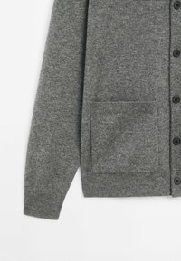 Manche d'un cardigan gris en tricot avec poche appliquée à l'avant et boutons noirs sur le côté droit, présenté sur fond blanc.