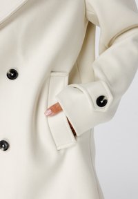 Hand met roze manicure in de zak van een crèmekleurige wollen jas met zwarte knopen en manchetstrapdetails tegen een lichte achtergrond.